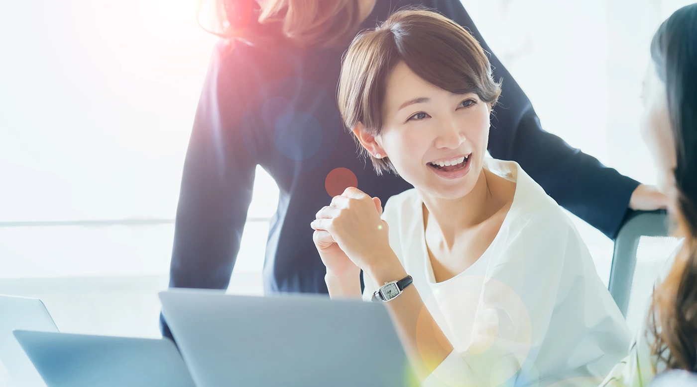 パソコンの前で同僚と笑顔で話す女性の公式ページ写真